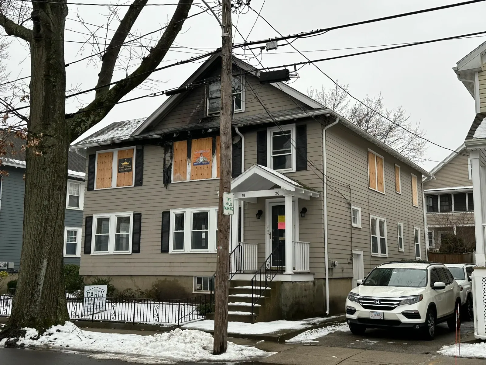 A house with boarded-up windows, black shutters, and a small front porch with stairs, located on a snowy street with parked cars and a utility pole with power lines.
