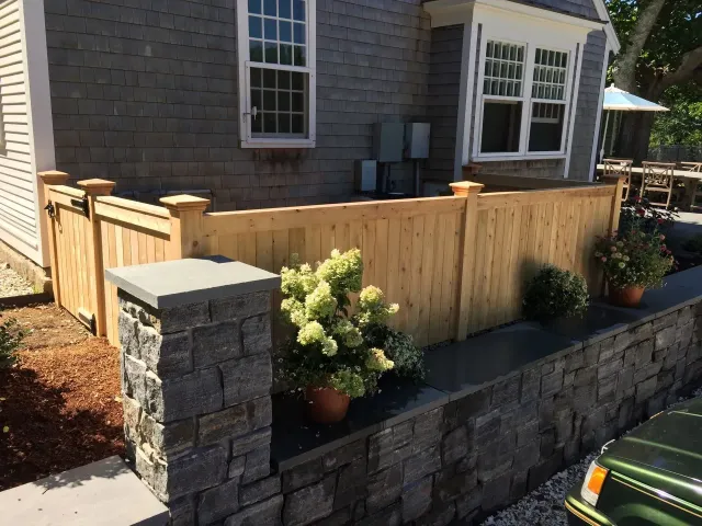 A wooden privacy fence built atop a gray stone wall, with potted plants on the ledge and a small gate near a house exterior.