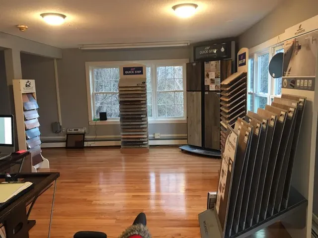 Interior of flooring showroom with hardwood floors, display racks of flooring samples, and a window.