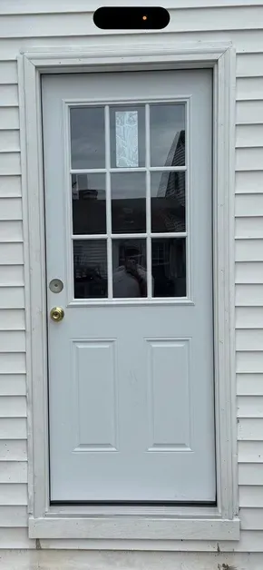 A white, nine-pane glass panel door installed in light gray vinyl siding with a brass-colored round doorknob.