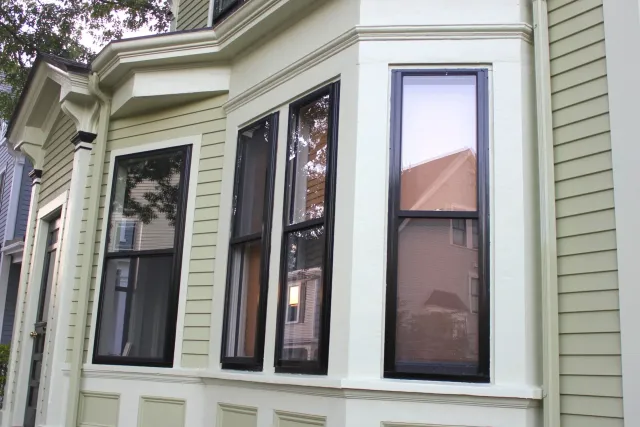 Close-up of a house's exterior  bay windows and green siding, black trim around windows, and decorative molding below the windows on Custer Street, Jamaica Plain.