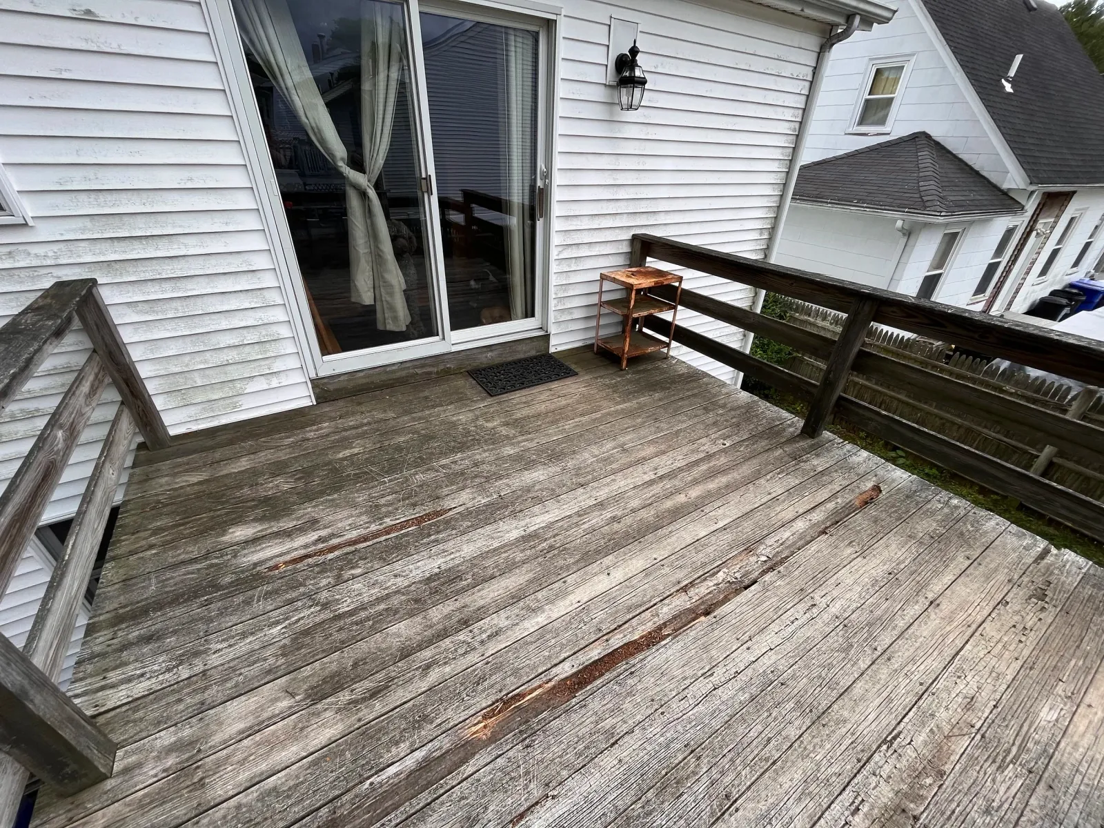 A weathered wooden deck attached to a white house with sliding glass doors and beige curtains. A small wooden shelf stands near the door. The house has white siding, a black exterior light, and a black rubber doormat.