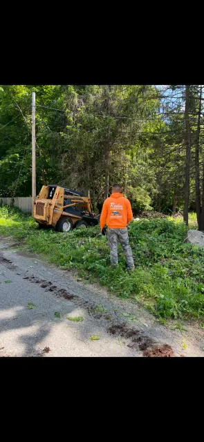 A man in an orange shirt is standing next to a tree stump grinder.