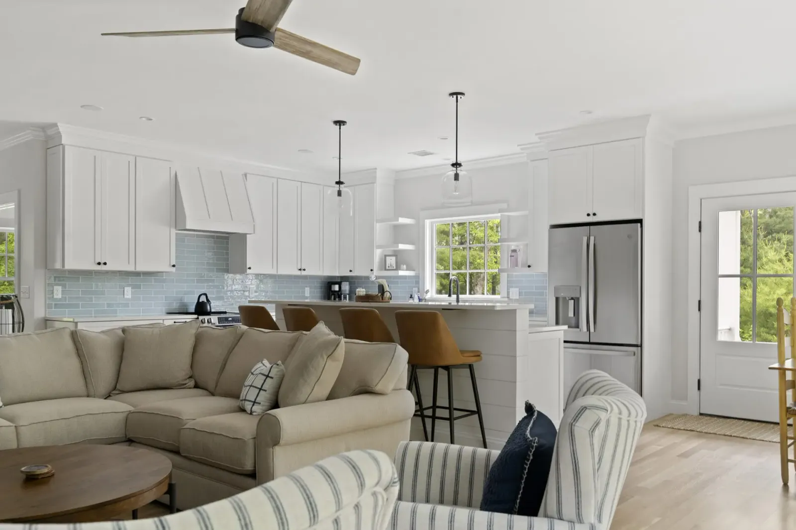 Modern open-concept kitchen and living room with white cabinetry, light blue subway tile backsplash, stainless steel appliances, beige sectional sofa, striped accent chairs, and wooden ceiling fan.