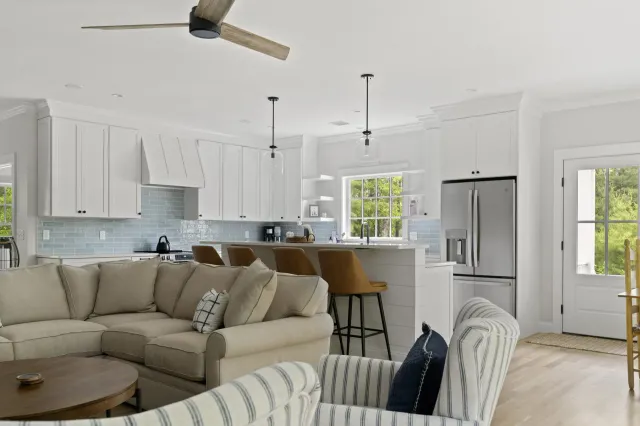 Modern open-concept kitchen and living room with white cabinetry, light blue subway tile backsplash, stainless steel appliances, beige sectional sofa, striped accent chairs, and wooden ceiling fan.