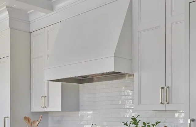 Close-up of a white kitchen range hood above a tiled white kitchen backsplash with white cabinets and silver handles.