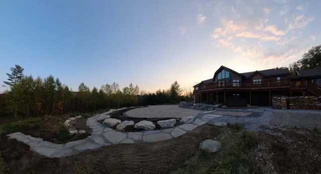 A wide-angle view of a rustic wooden house at dusk, featuring a stone patio and a rock-bordered path in the foreground.