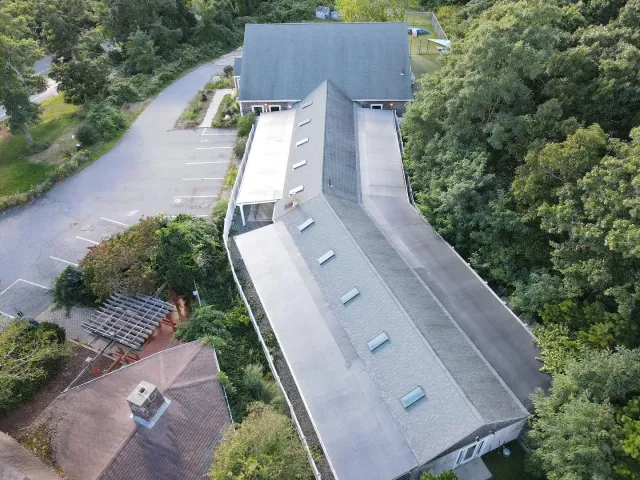 Aerial view of a curved gray-roofed building beside a parking lot and trees