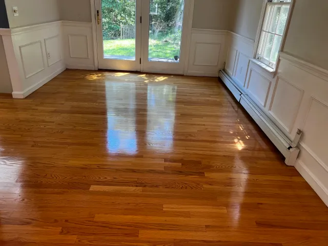 Empty room with shiny hardwood floor, white wainscoting, and large windows and glass door leading to a backyard with green grass and trees.
