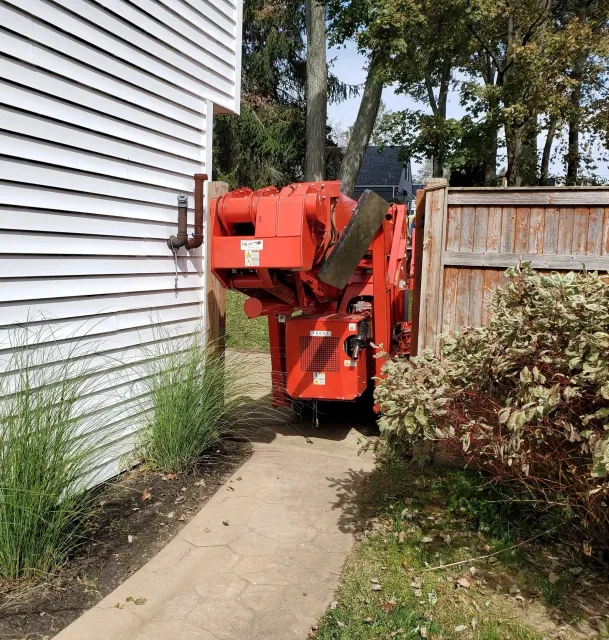 A sidewalk runs between a white house with siding and a garden bed with grass and plants on the left, and a wooden fence with bushes on the right. A red utility lift is parked in a yard behind the sidewalk, with trees and a house visible in the background.