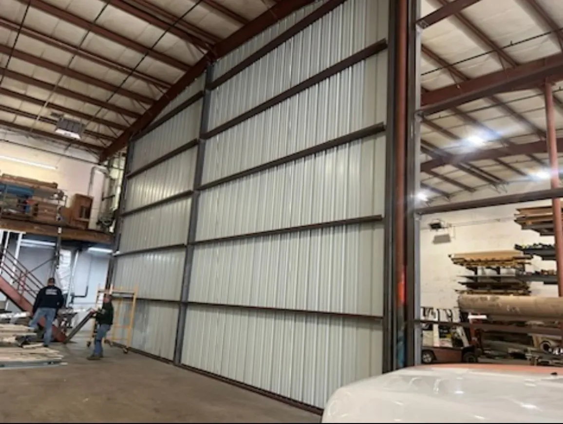 Interior of a metal workshop with a tall sliding door, steel beams, and workers near equipment