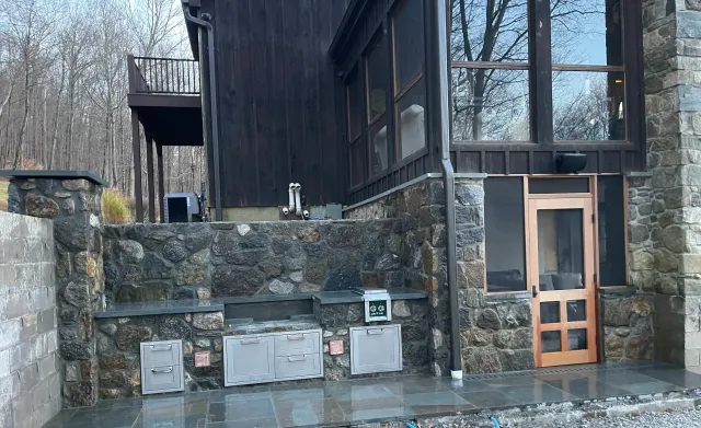Outdoor kitchen area with stone counter, cabinets, and a wooden door, adjacent to a dark wood building with large windows, surrounded by a stone patio and trees in the background.