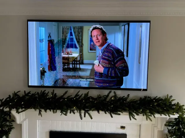 Man in a striped sweater holding a mug in a living room, shown on a wall-mounted TV.