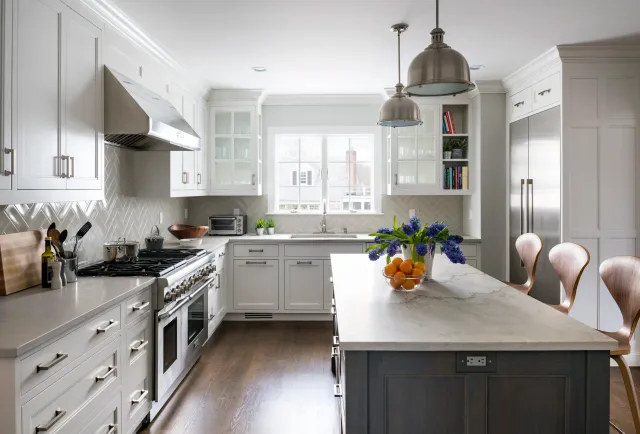 Modern white kitchen with an island, wooden chairs, stainless steel appliances, a window above the sink, and decorative vases with flowers and fruit on the island.