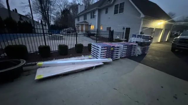 Long white board lying on driveway beside stacked lumber and a fenced house at dusk