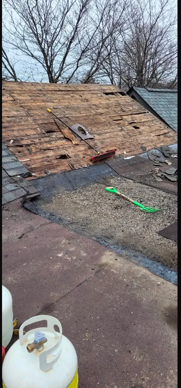 Damaged roof with exposed plywood, scattered debris, and a propane tank on a flat rooftop.
