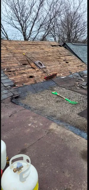 Damaged roof with exposed plywood, scattered debris, and a propane tank on a flat rooftop.