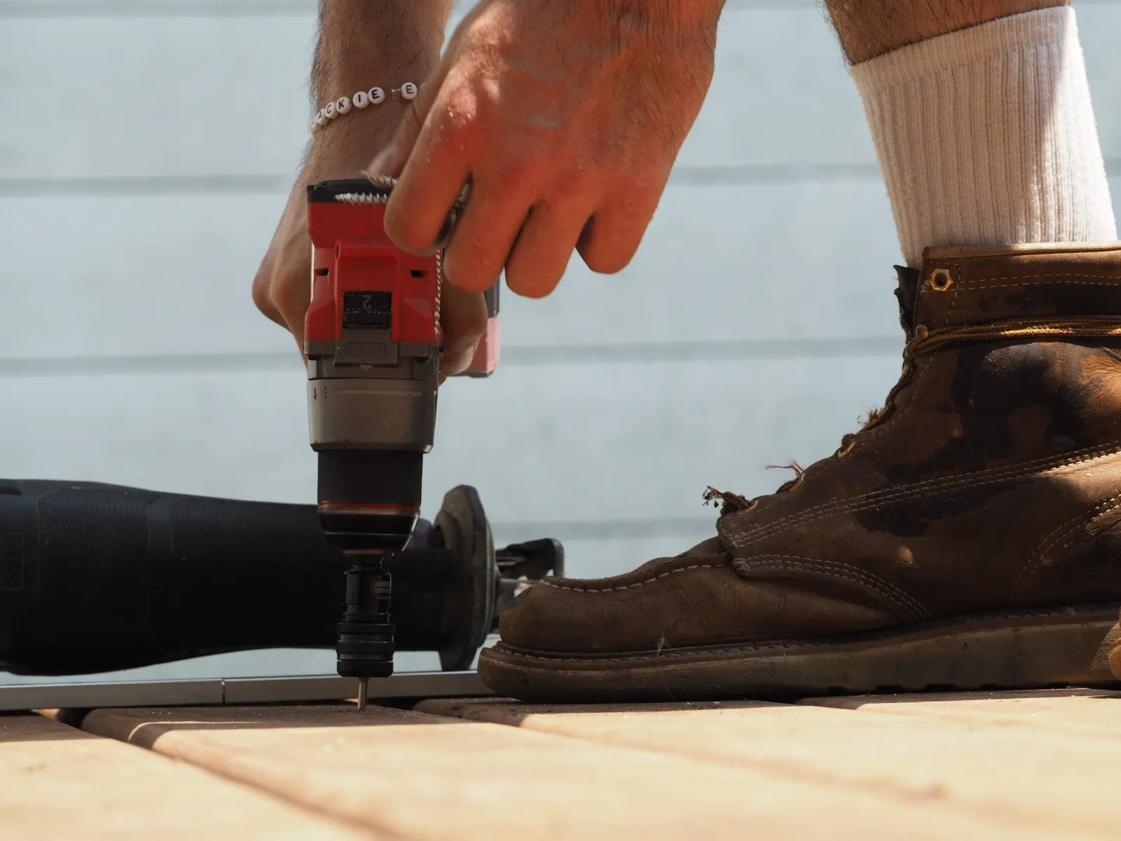 Close-up of a person's hand using a cordless drill on a wooden surface, with worn brown leather work boots and a white sock visible.