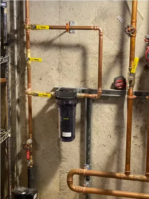 Basement utility wall with copper pipes and a mounted black water filter.