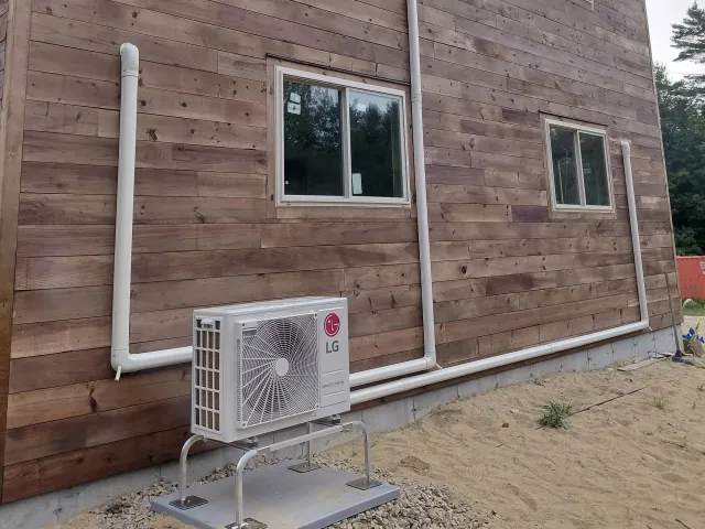 Exterior view of a wooden house with two white-framed windows. An LG heat pump is installed outside, connected with white pipes running along the wall. The ground is sandy with some grass patches.
