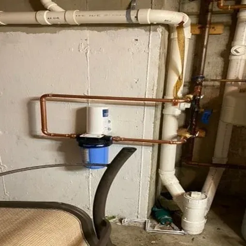A water filter system with copper pipes is mounted on a basement wall next to white PVC plumbing. Part of a chair is visible in the lower left corner of the image.