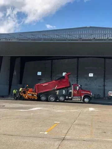 Red dump truck unloading asphalt into a paving machine near a large gray building.