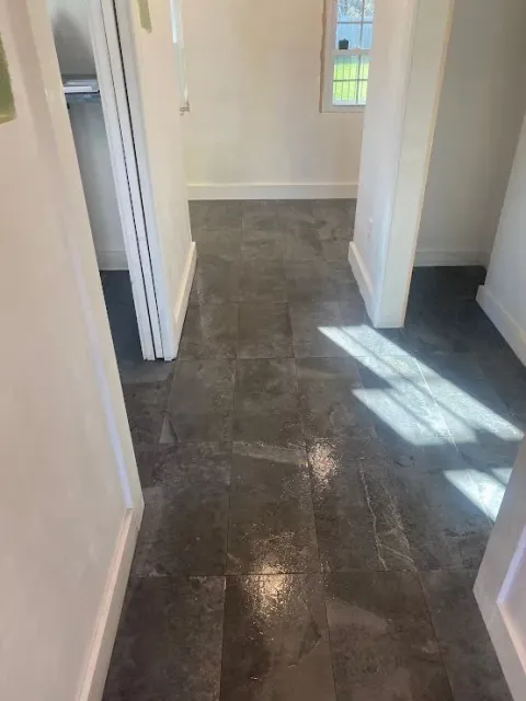 A high-angle view of a hallway featuring dark grey, textured stone tile flooring with bright sunlight casting shadows.