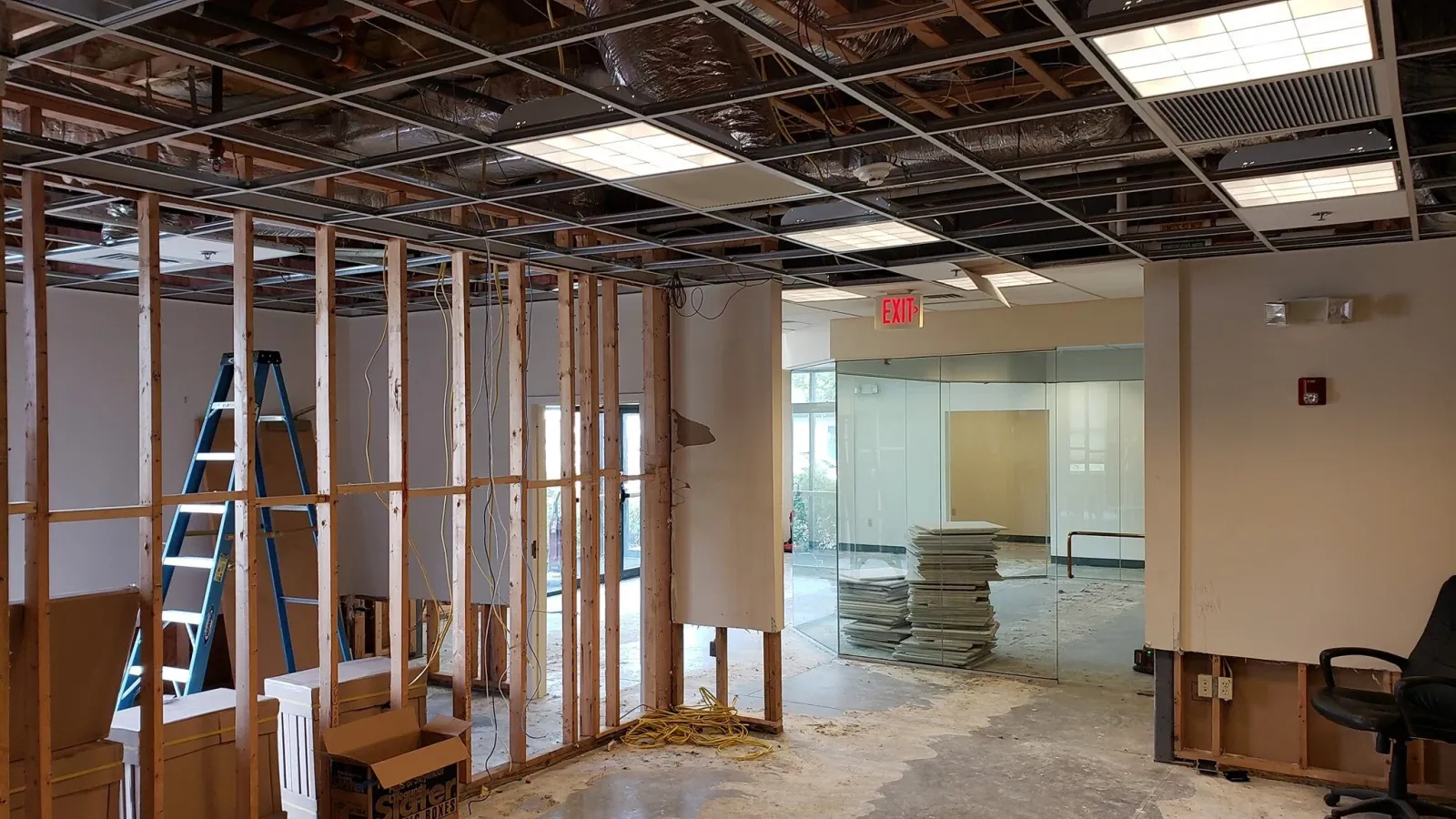 Interior of a building under renovation with exposed ceiling framework, a ladder, construction materials, and a partially open wall.