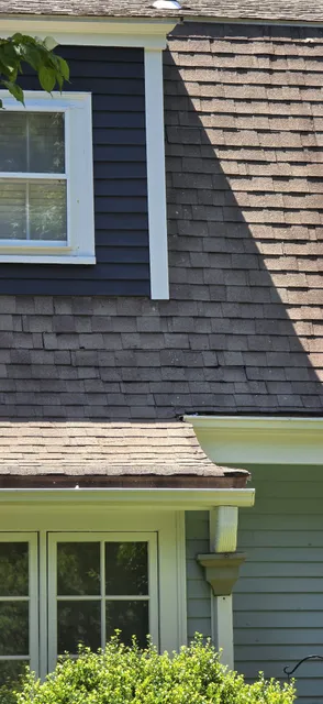 Detailed view of a residential house exterior, showcasing dark blue siding, aged asphalt shingle roof, and a gutter system that may need attention in Wellesley. Contact Handyman Boston for reliable home services.