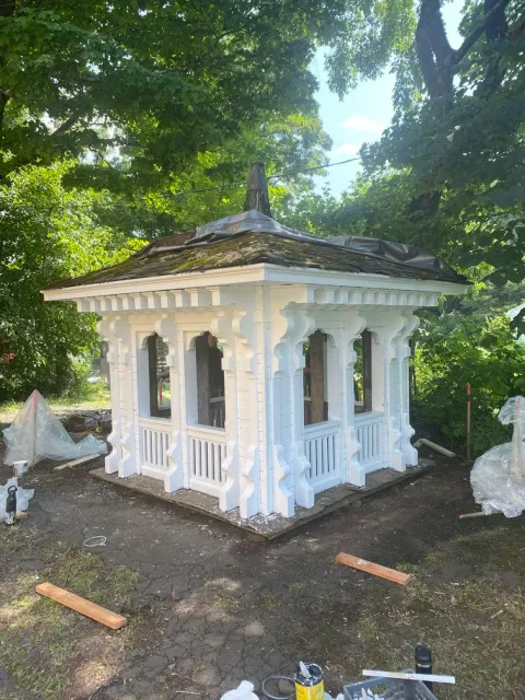 A small, decorative white gazebo with intricate architectural details, situated outdoors among green trees, with construction materials and tools around it.