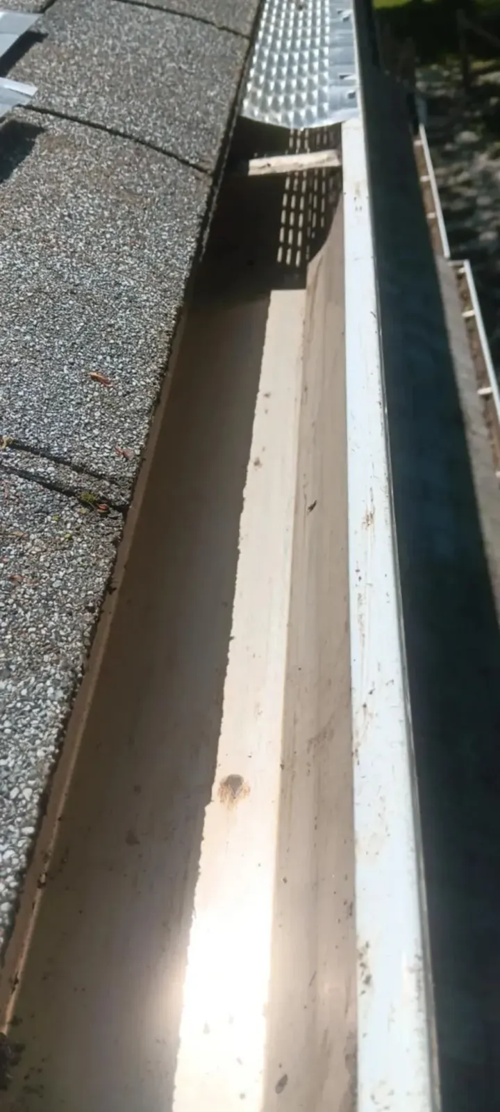 Narrow rooftop gutter beside shingles, with bright sunlight and shadows.