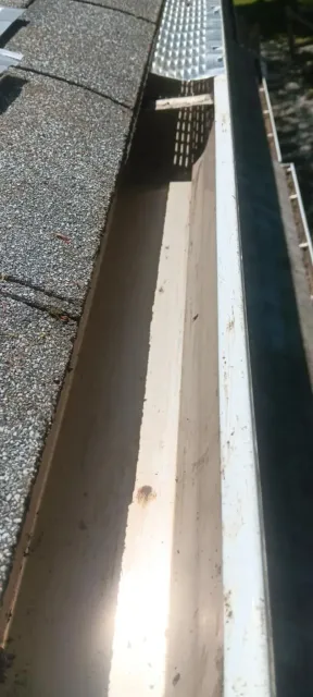 Narrow rooftop gutter beside shingles, with bright sunlight and shadows.