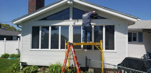 Person standing on scaffolding working on a white house's exterior trim under a triangular window.