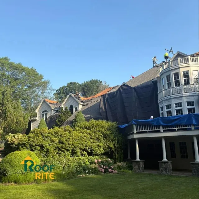 Roof repair on a large house with a tarp and workers. Green yard with trees and blue sky.
