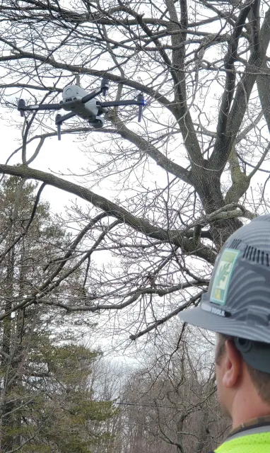A person wearing a baseball cap with a sticker and safety vest, observing a drone flying near leafless tree branches during a cloudy day.