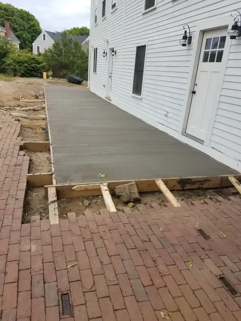 New concrete patio next to a white building. Red brick pathway on the left.