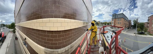 A worker in a yellow hazmat suit uses a power washer on the brick exterior of a building while standing in a lift.