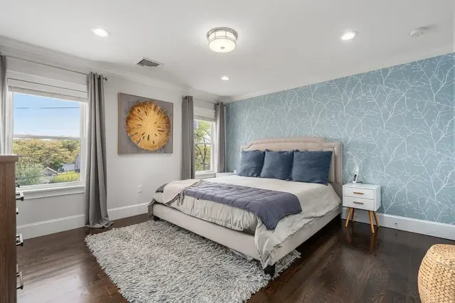 A bedroom with a large bed with a beige headboard, dark blue pillows, and a gray bedspread. The room has two windows with gray curtains, a white nightstand, and a textured light blue wallpaper accent wall. There is a fluffy beige rug on the dark wooden floor.
