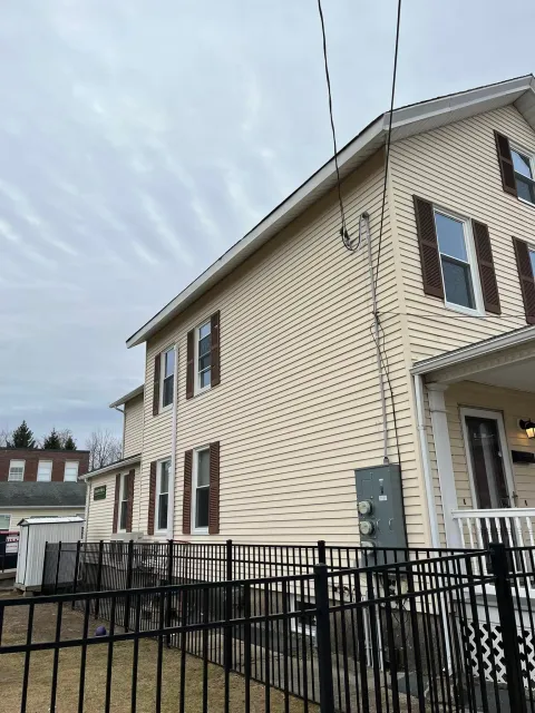 Residential house with beige siding, dark shutters, and a black metal fence under a cloudy sky