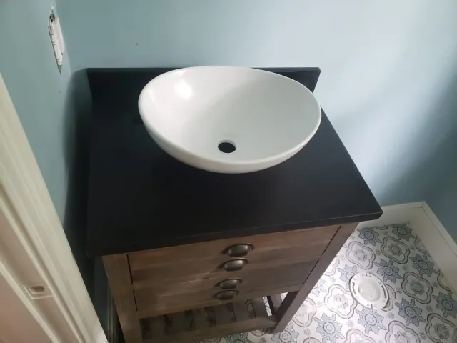 Bathroom vanity with a white bowl sink and dark countertop. Wooden drawers and a patterned floor.