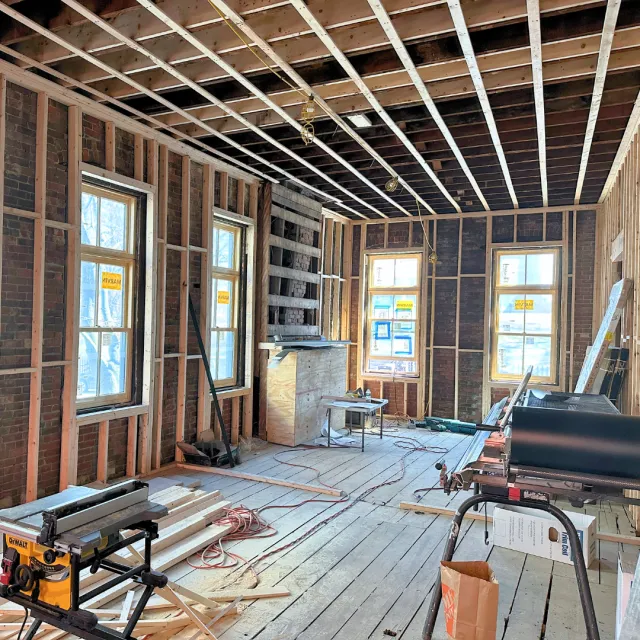 Room under construction with exposed wooden framing, three windows, and construction tools and materials on the floor.