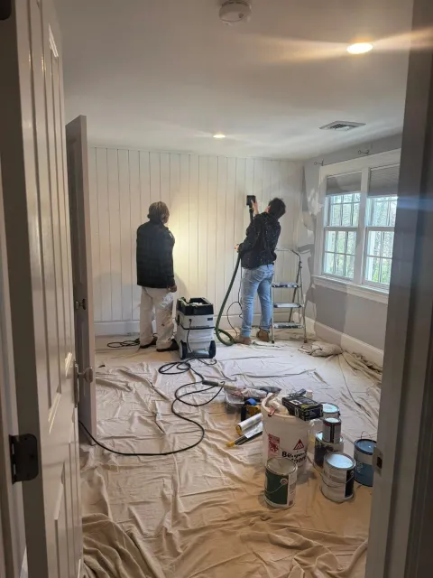 Two workers painting a room during renovation, with tools, drop cloths, and supplies scattered on the floor.
