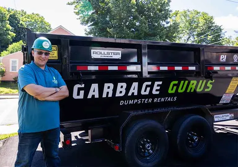 Man in blue shirt and green hard hat standing confidently next to Garbage Gurus dumpster rental truck, showcasing eco-friendly junk removal services.