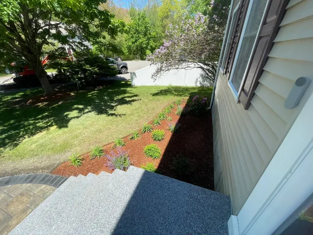 Front porch beside beige house, with a sloped lawn, flower bed, and driveway in bright sunlight.