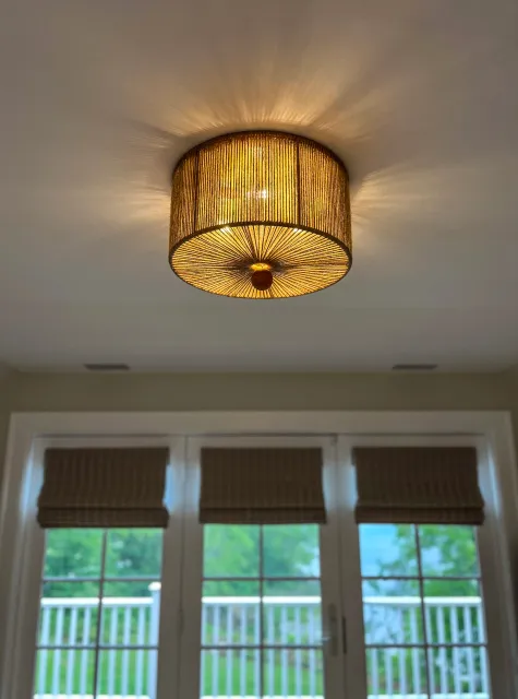 Ceiling light fixture with a woven rattan shade illuminating the room, ceiling with two small vents, and French doors with blinds leading outside to a porch with a white railing and green trees in the background.