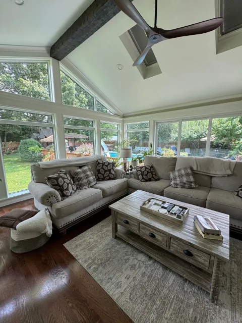 Bright sunroom with large windows, gray sofas, a wood coffee table, rug, and a ceiling fan over dark hardwood floors