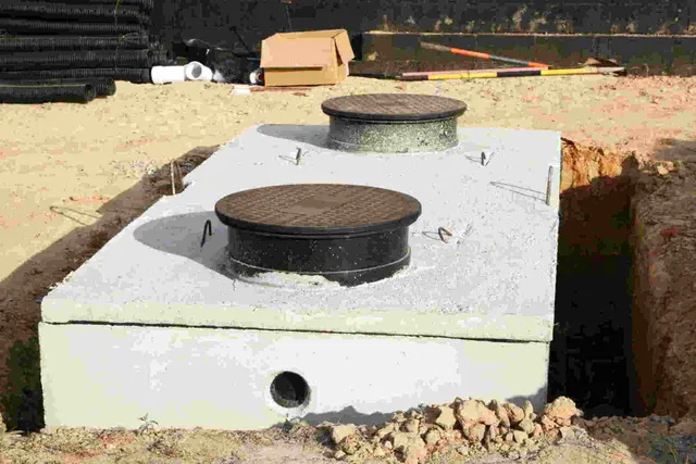 Two round manhole covers on a white concrete septic tank set in sandy ground.