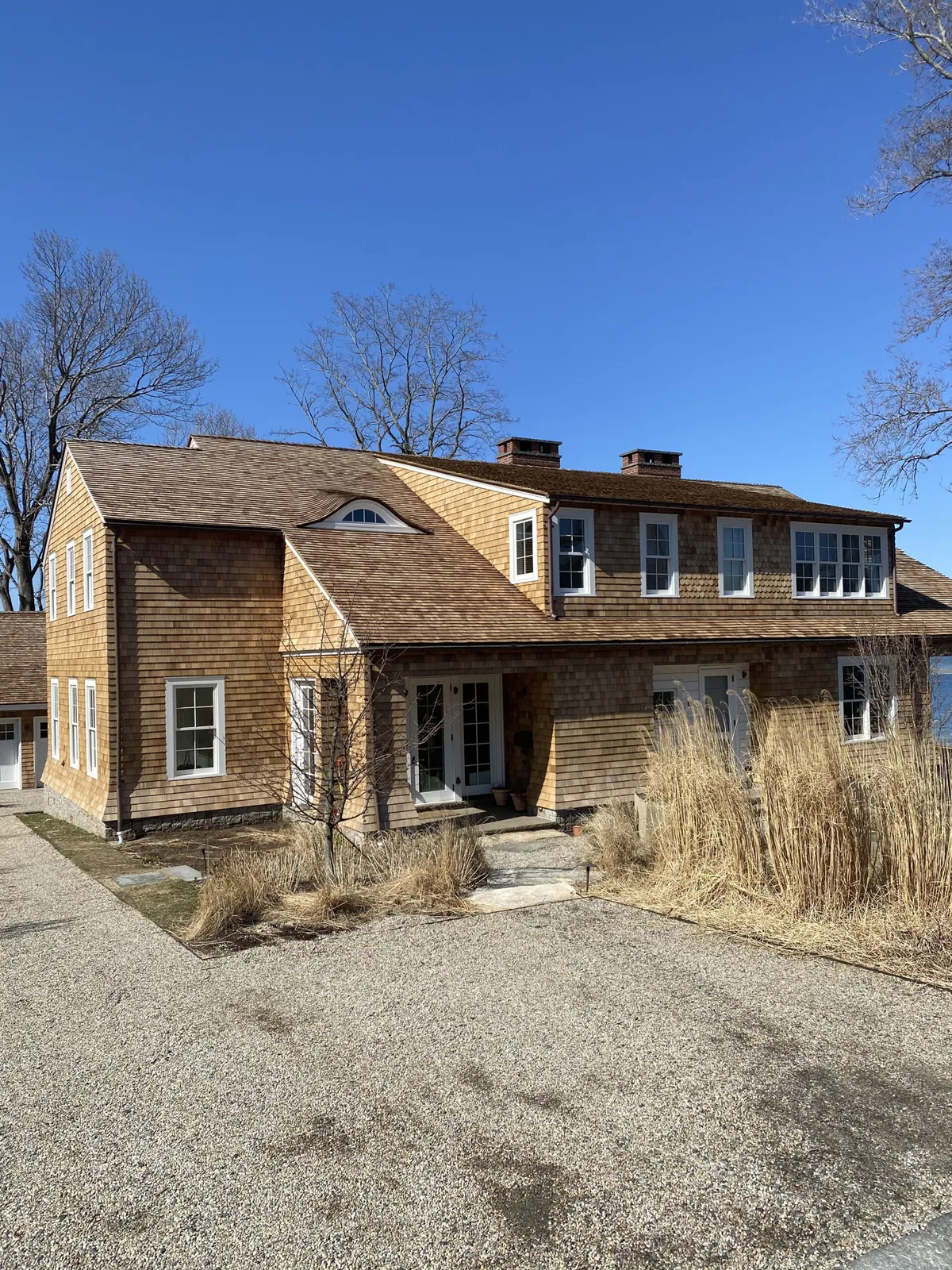 A large two-story house with brown shingles, white trim, and a covered front porch. Tall dry grass lines the pathway, and leafless trees stand in the background under a clear blue sky.