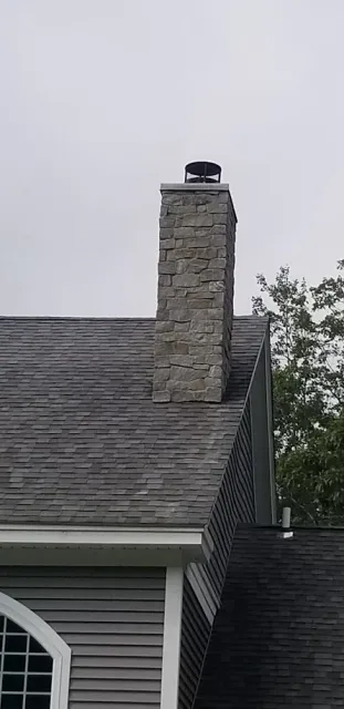A stone chimney on the roof of a house with gray siding and a gray shingle roof, overcast sky in the background.