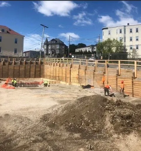 Construction workers building a wooden retaining wall at a construction site in an urban area with multi-story buildings and power lines in the background.